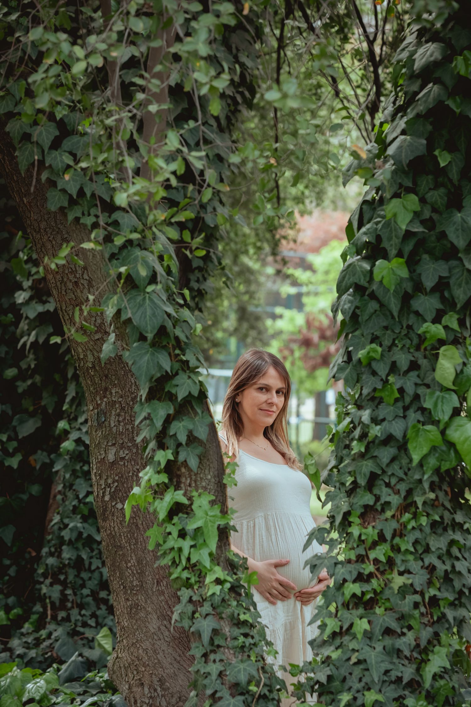 Pregnant woman in white dress standing between ivy-covered trees in a serene green park.