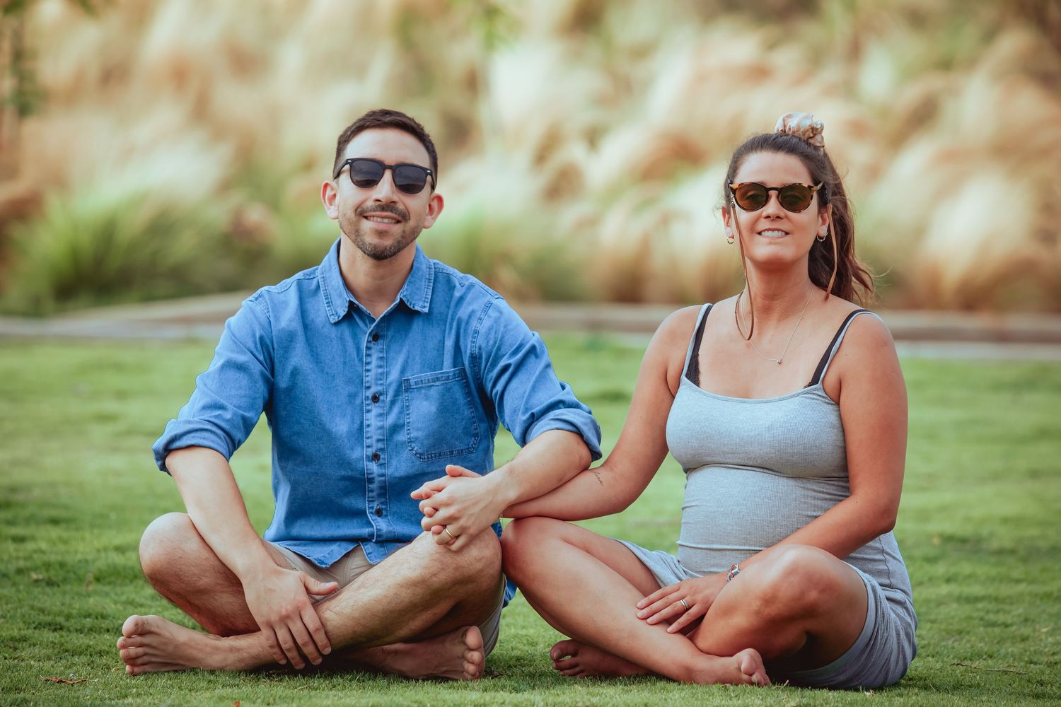 Happy couple sitting cross-legged on green grass, both wearing sunglasses, holding hands outdoors.
