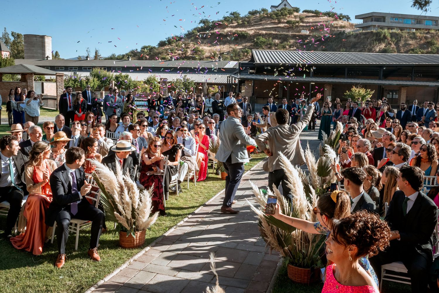 Wedding couple walks down the aisle as guests celebrate with confetti at a sunny outdoor ceremony.