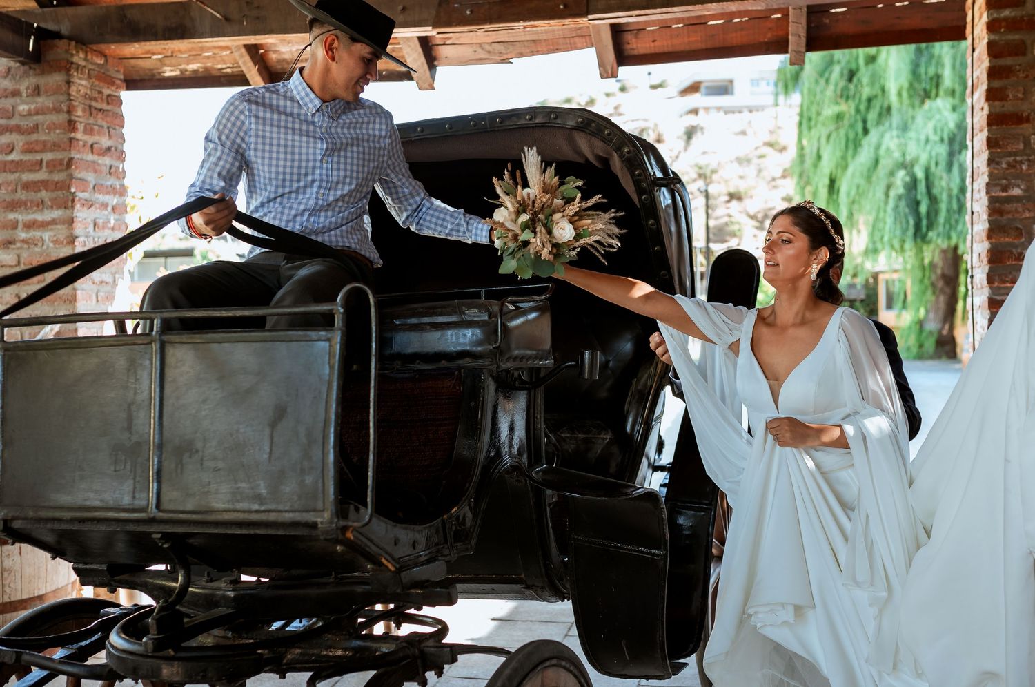 Bride in white gown handing her bouquet to a coachman seated on a vintage black horse-drawn carriage.