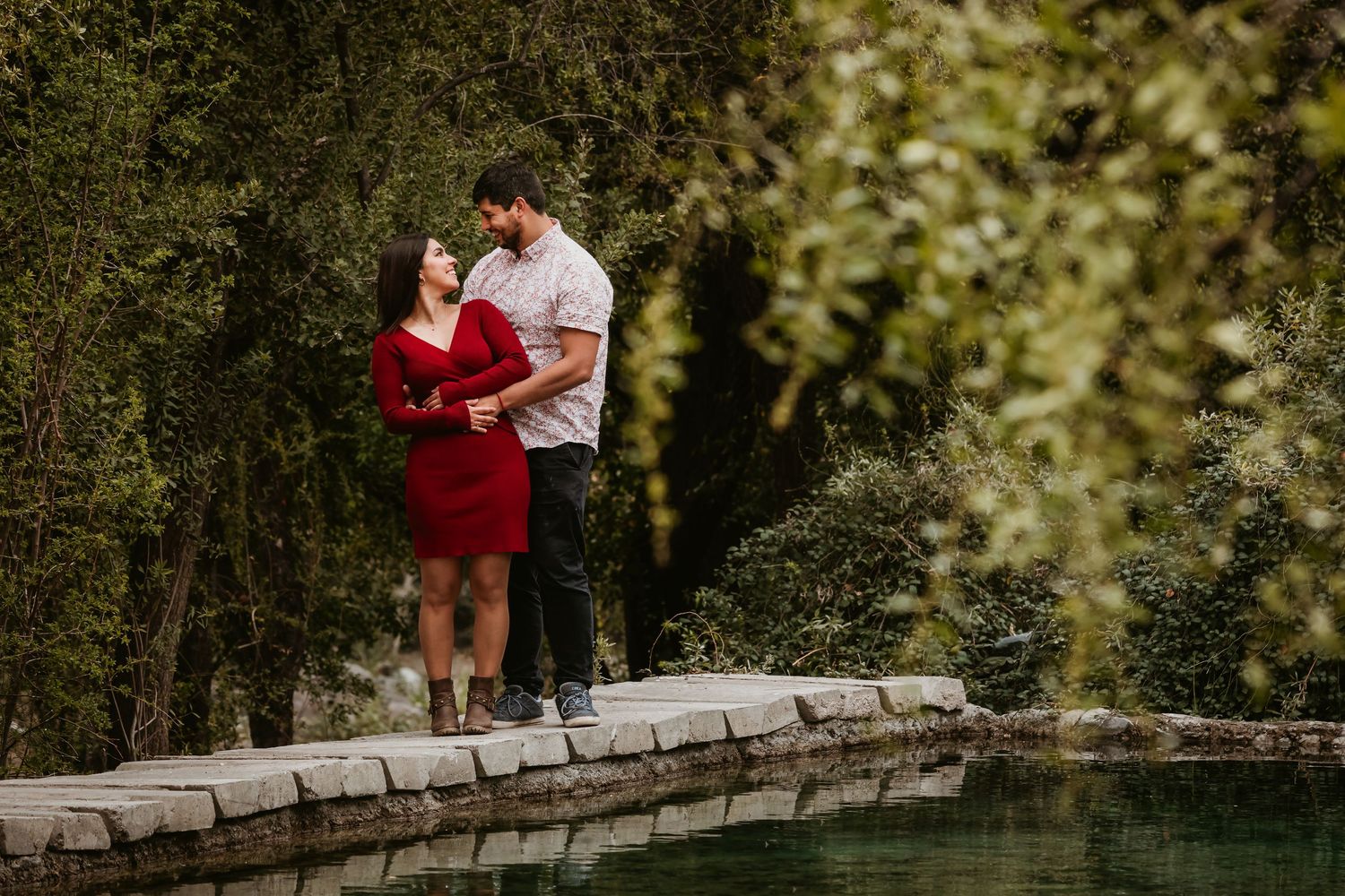 Couple standing close together on a stone path beside a calm green reflecting pond.