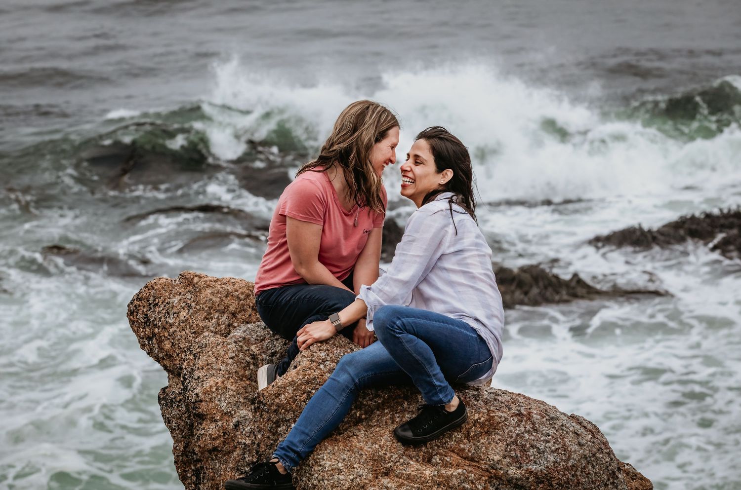 Two women sitting close on coastal rocks with crashing ocean waves behind them.