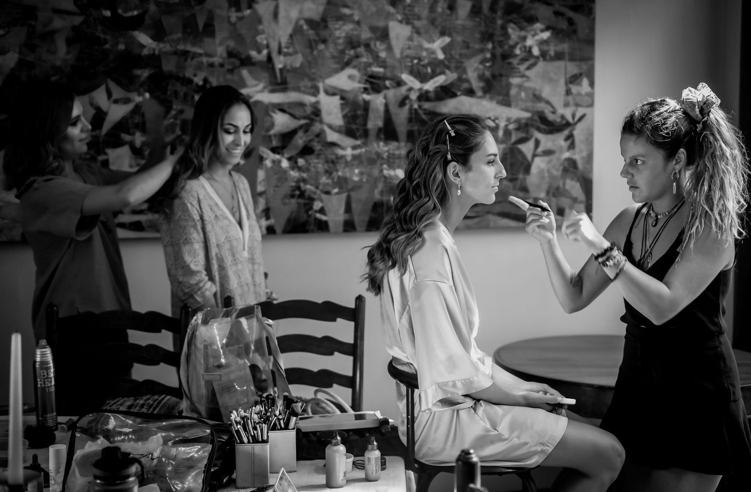 Black and white photo of bride in white robe getting hair and makeup done by two stylists before wedding.