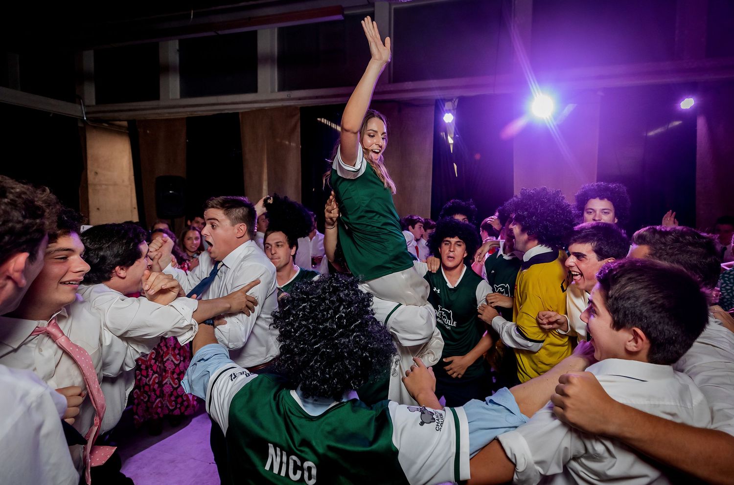 Excited wedding guests lift a person in a green jersey during a lively celebration on the dance floor.