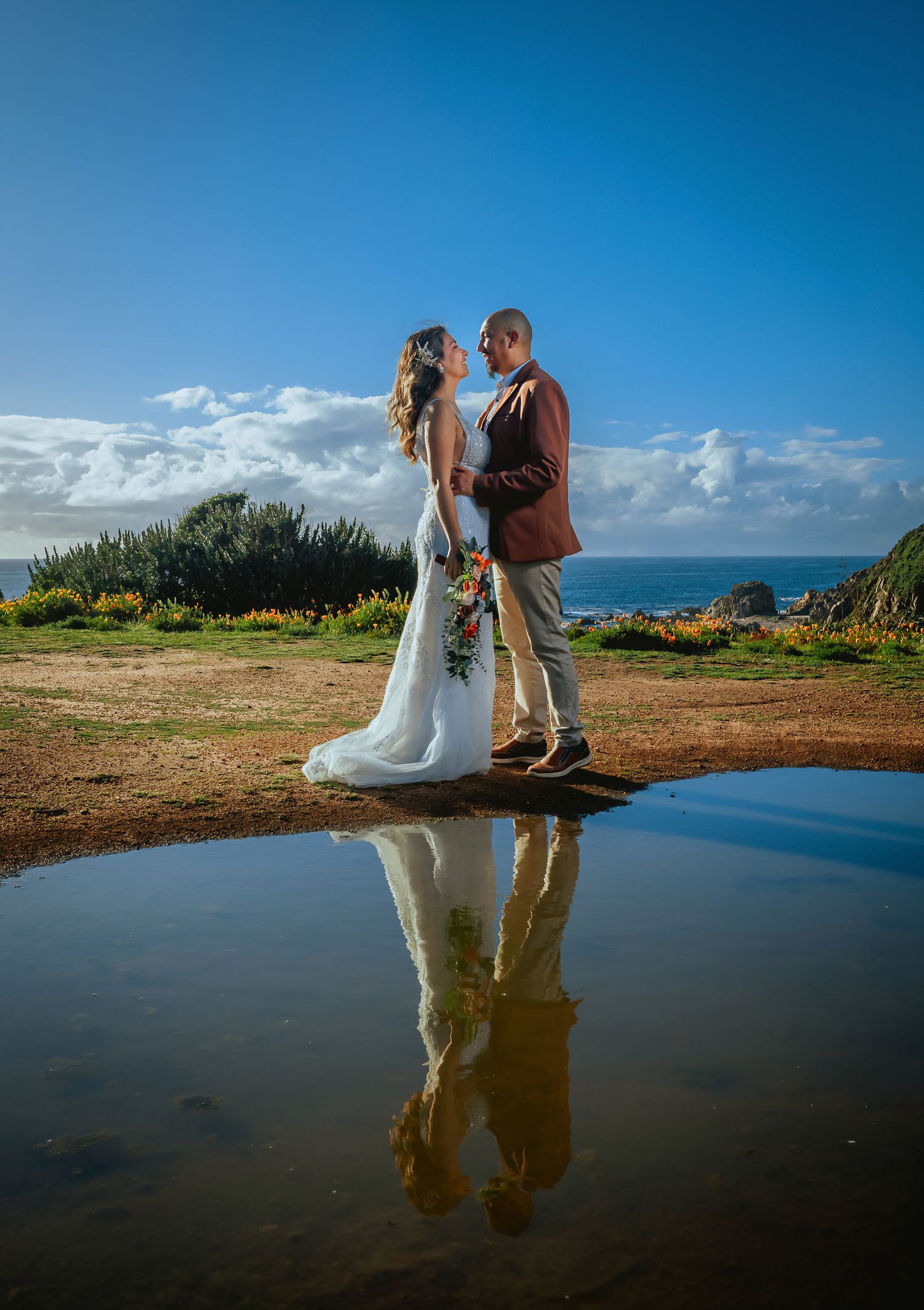 Bride in a white gown and groom in a brown blazer embrace on a coastal bluff with ocean views, their reflection mirrored in a puddle.