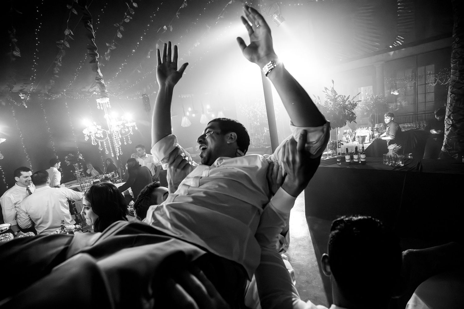 Black and white photo of a man raising his hands at a lively party or wedding reception celebration.