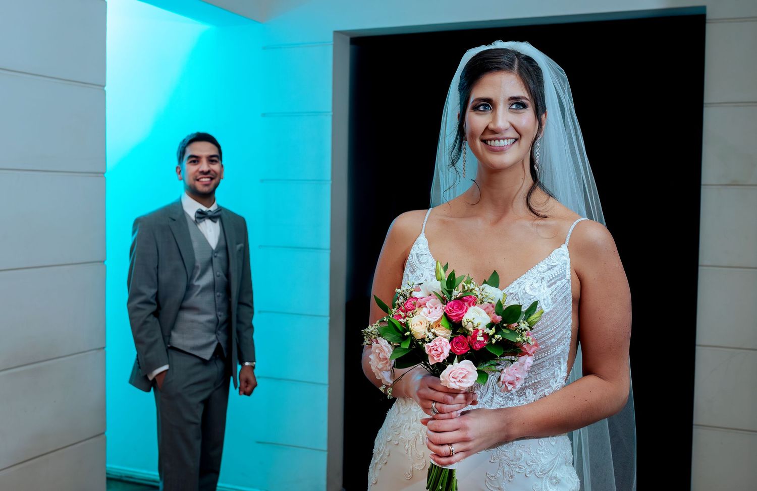 Bride in white lace gown holding floral bouquet with groom in gray suit standing in elegant wedding venue.