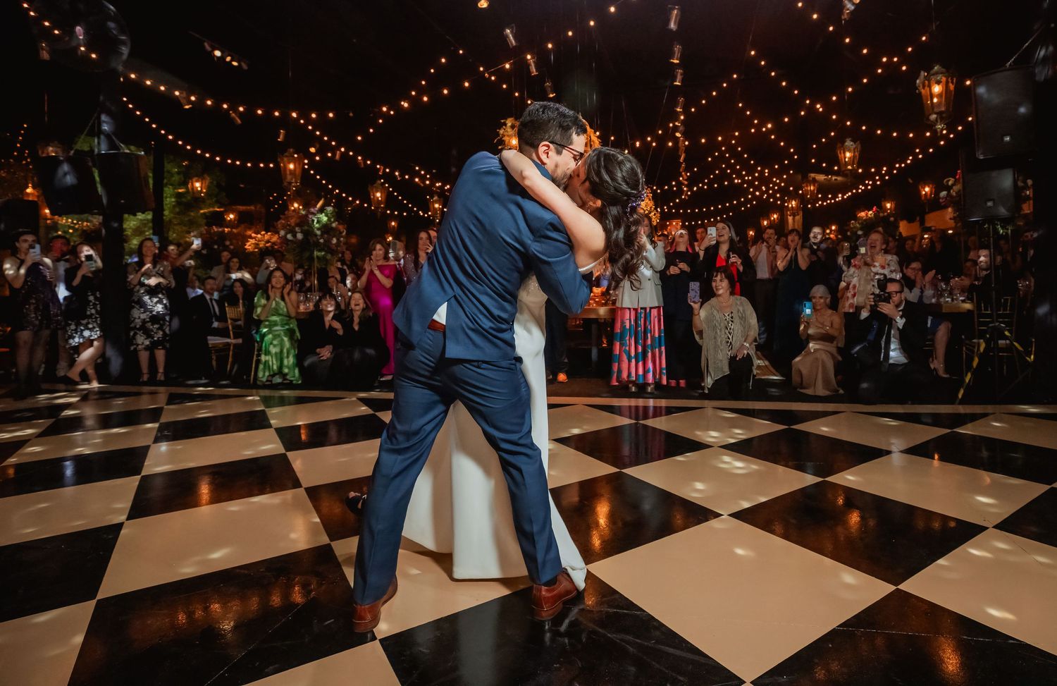 Couple shares romantic first dance embrace on checkered ballroom floor at elegant wedding reception with string lights.