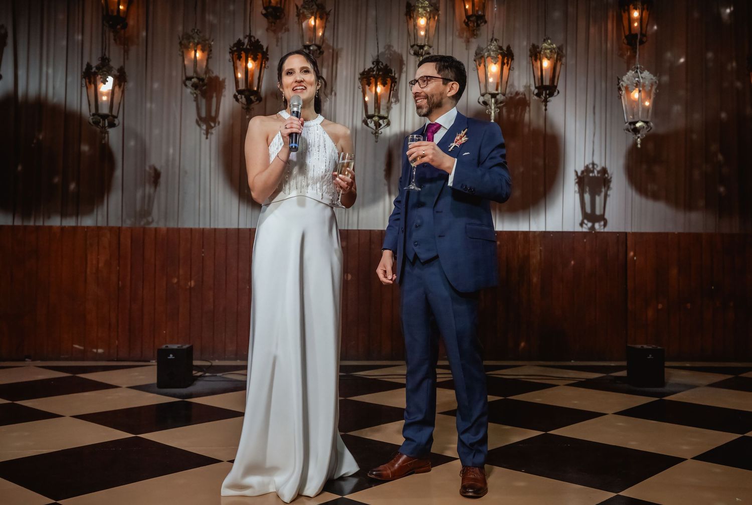 Bride and groom giving a toast speech together on a checkered dance floor at their wedding reception.
