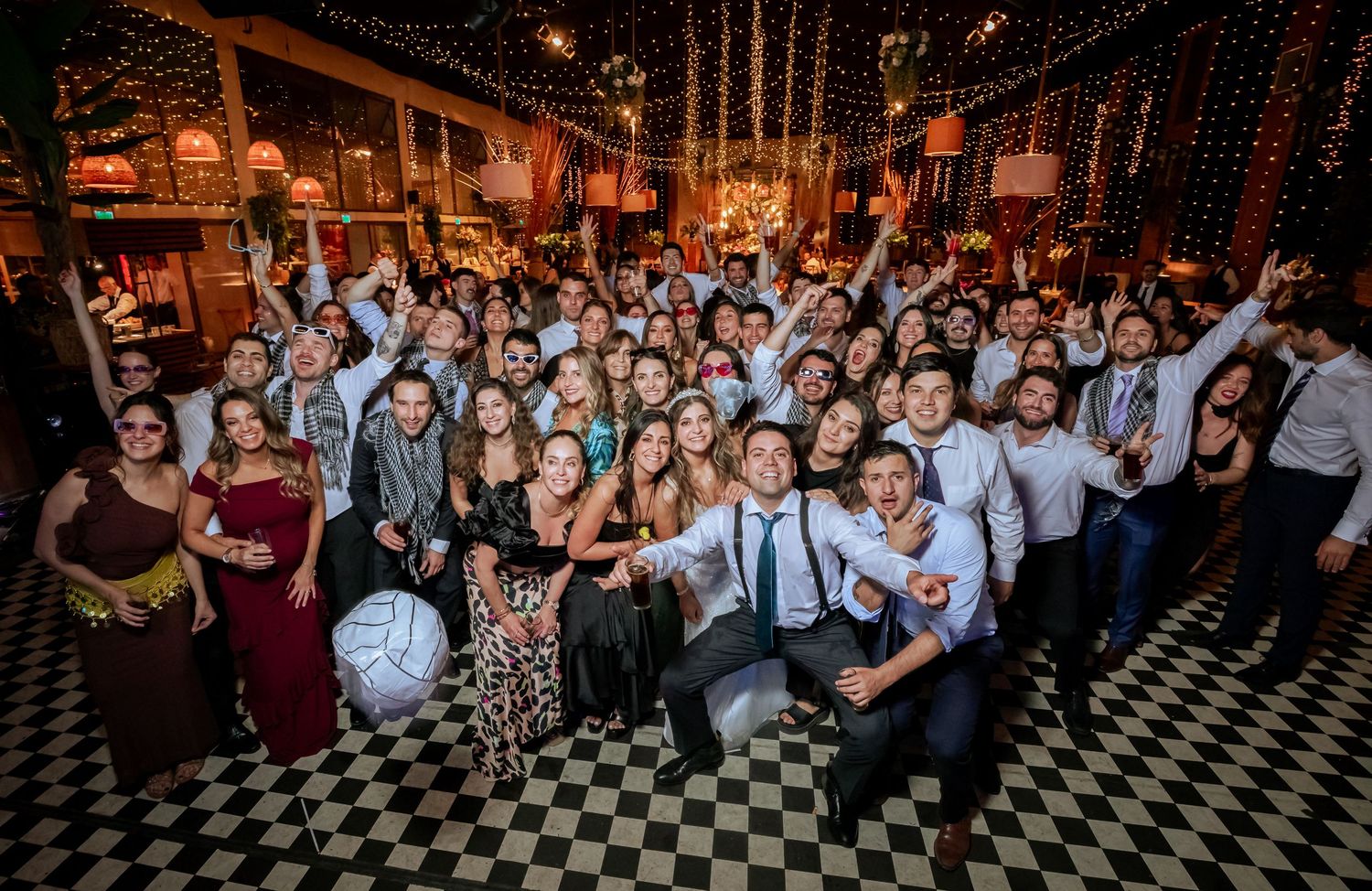 Large group of wedding guests posing together cheerfully on a checkered dance floor at reception.