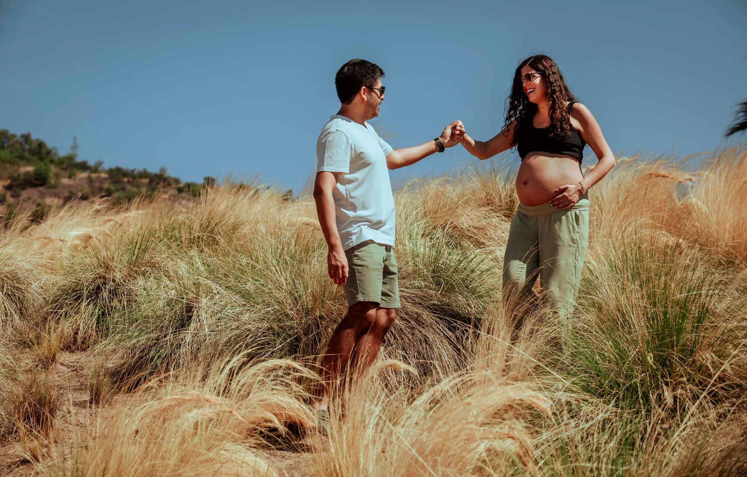 Couple facing each other in a golden grass field, man gently touching pregnant womans hand outdoors.