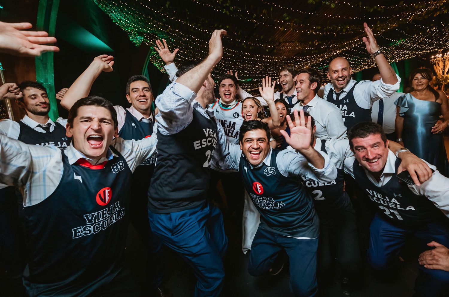 Large group of men in Vieja Escuela jerseys cheering and celebrating at a wedding.