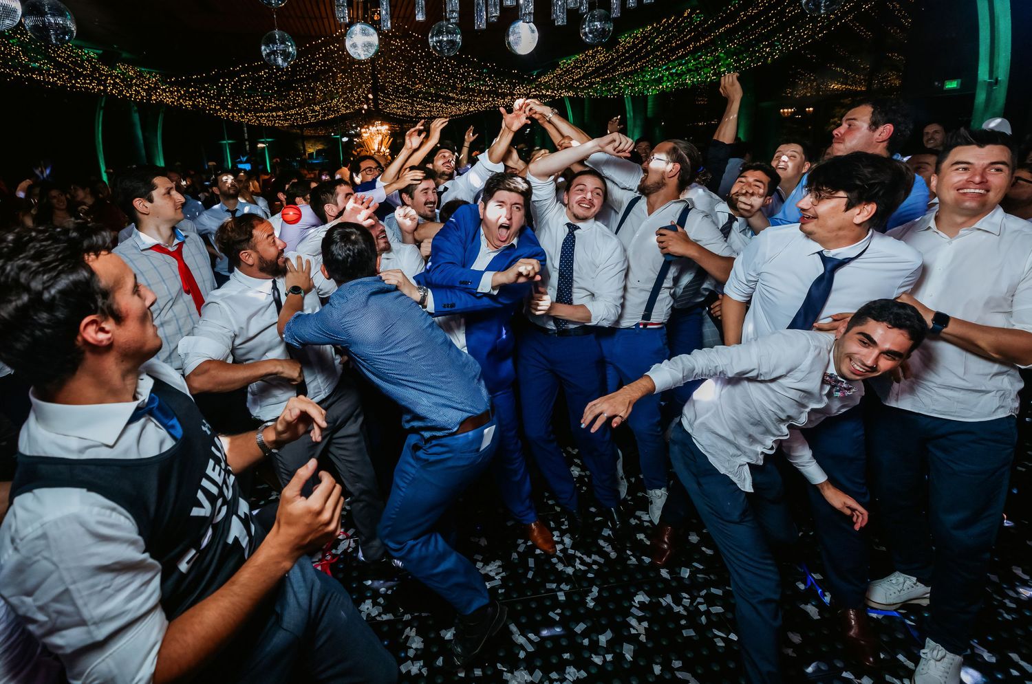 Groom in blue suit dancing energetically with guests at festive wedding reception.