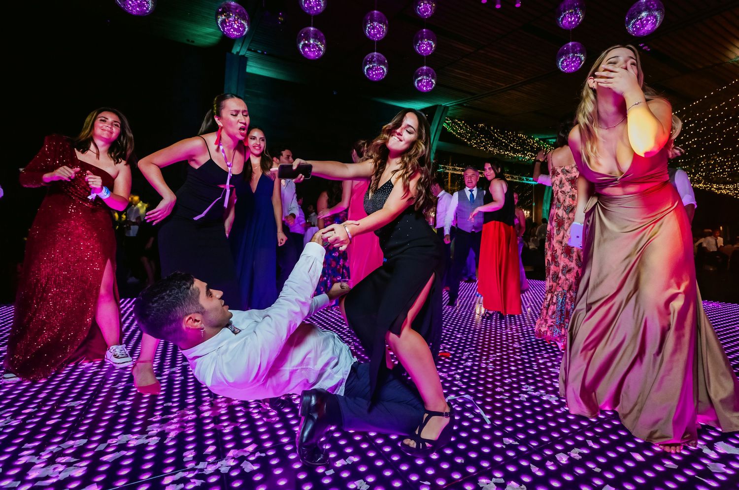 Wedding guests in colorful gowns dancing energetically on purple-lit dance floor.