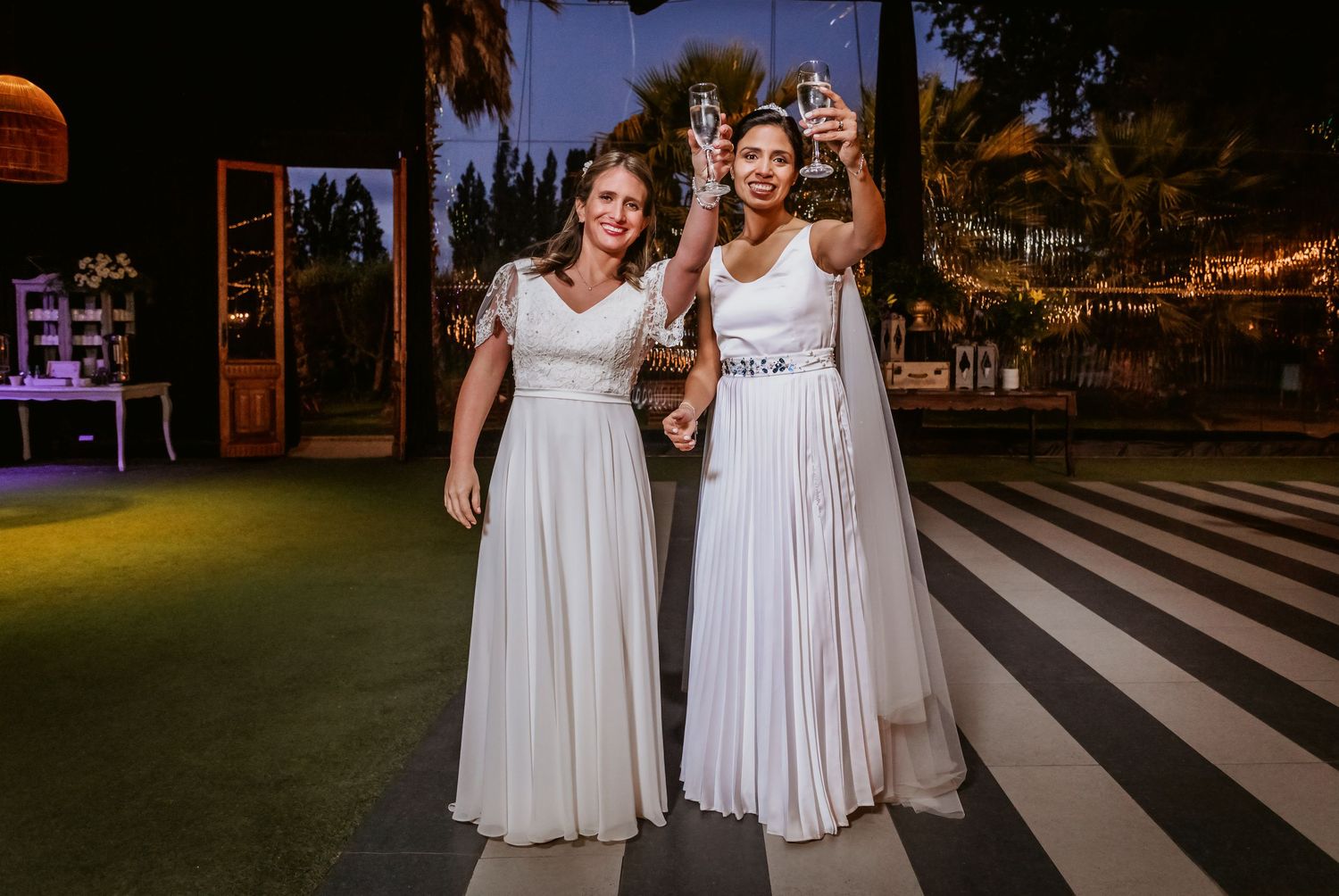 Two brides in white wedding gowns smiling on striped dance floor at outdoor wedding reception.