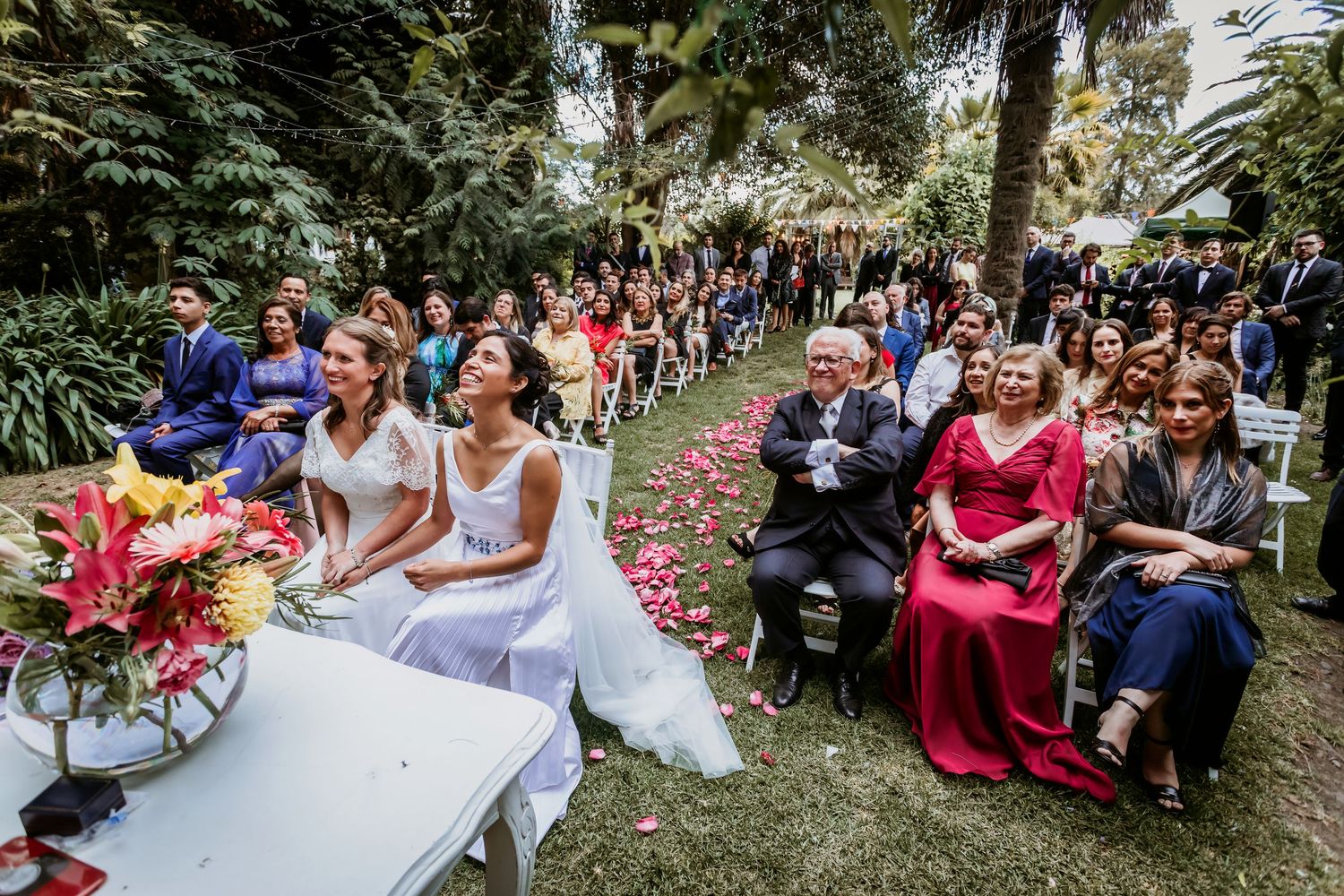 Two brides sit at ceremony table as guests and family watch outdoor garden wedding ceremony.