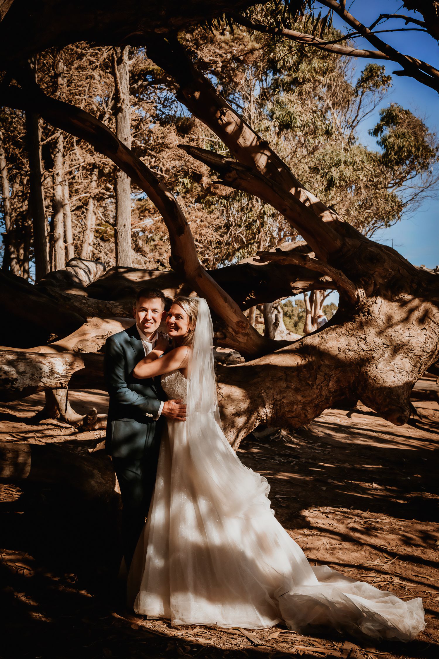 Joyful bride and groom laughing together beside large twisted tree roots.