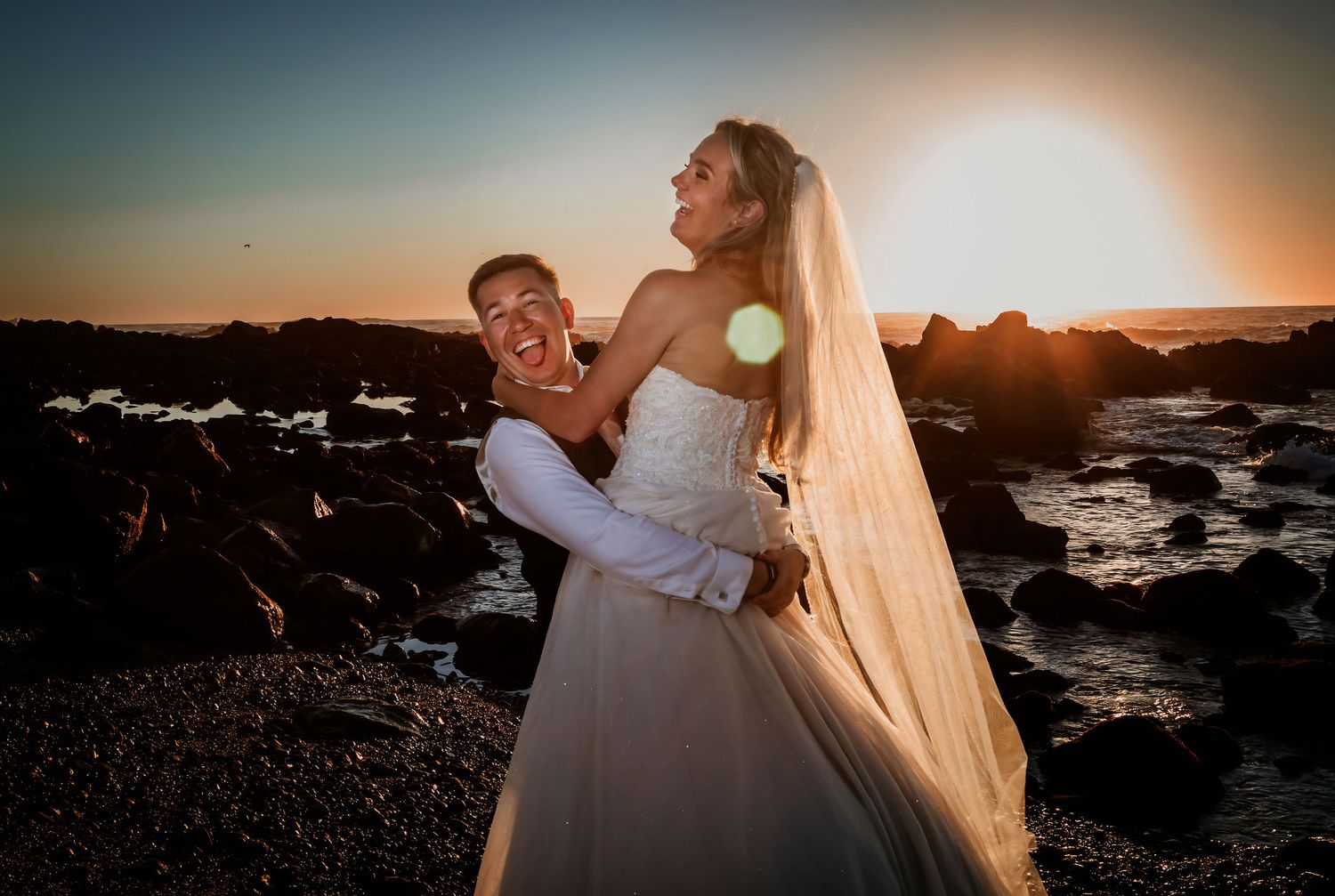 Laughing couple embracing on rocky beach at sunset, bride in strapless gown with long veil.