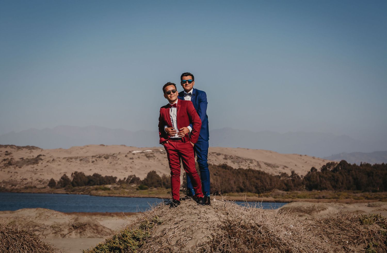Two grooms posing together in red and blue suits beside a tranquil lake with sand dunes behind them.