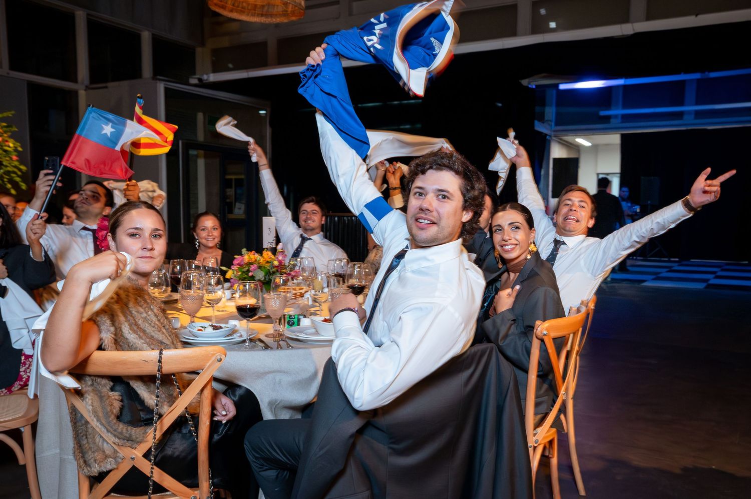 Excited wedding guests celebrating at dinner table waving Chilean flag in festive outdoor reception venue.
