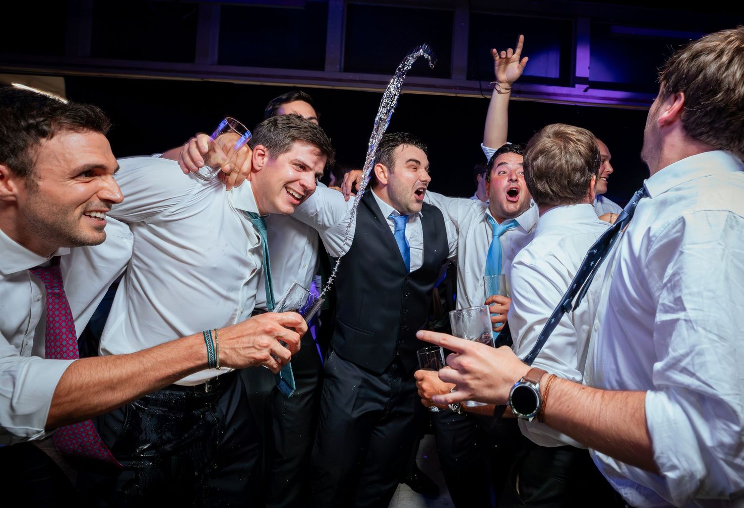 Excited groomsmen celebrating with drinks and dancing together at a lively indoor wedding reception party.