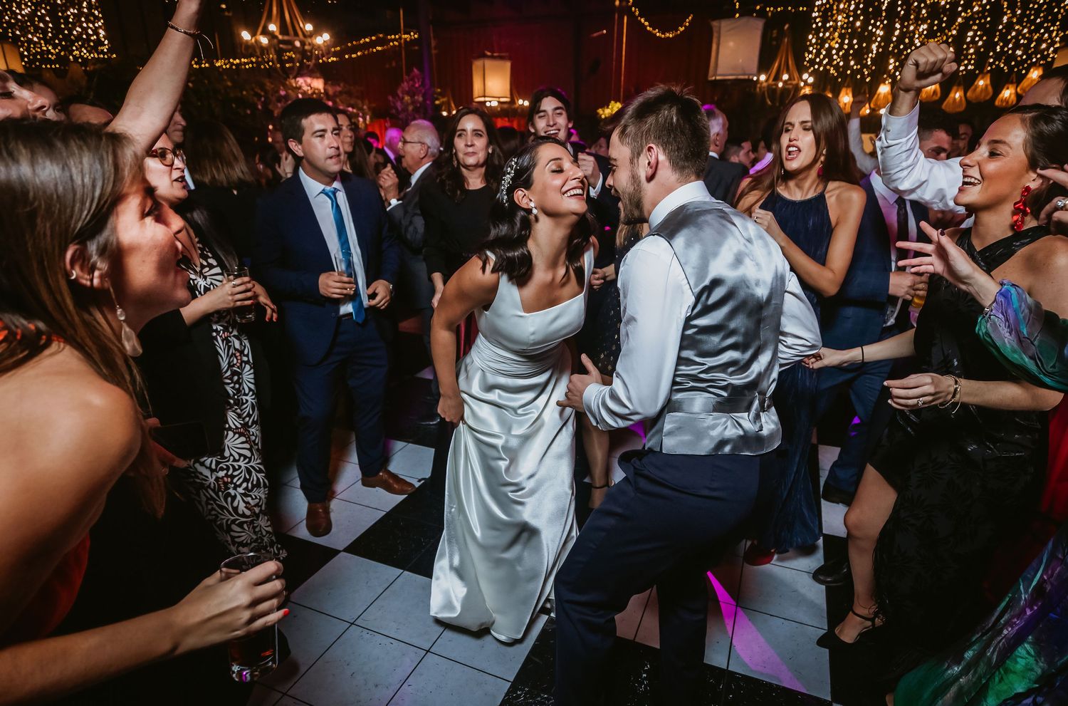 Bride and groom dancing joyfully with wedding guests on a festive dance floor with warm string lights.
