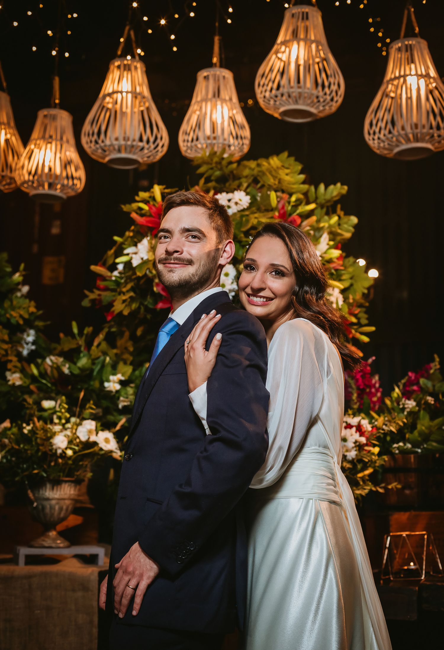 Bride and groom posing elegantly at wedding reception with warm pendant lights and lush floral backdrop.