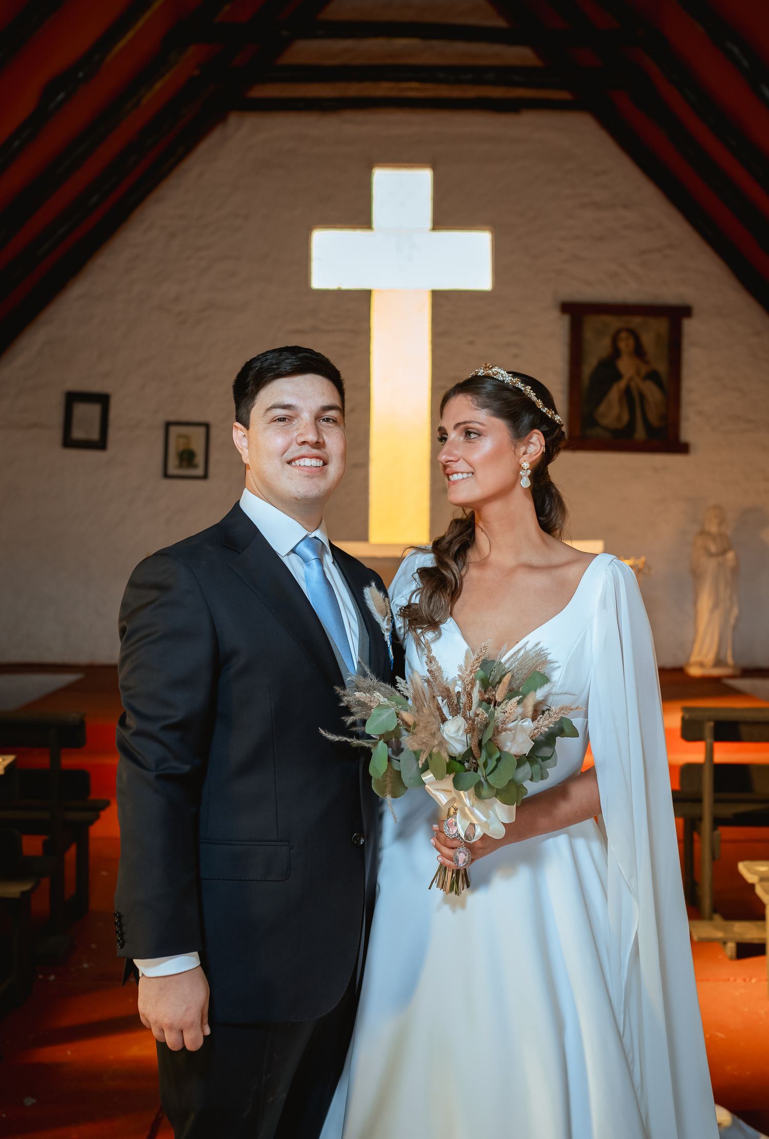Bride and groom posing together inside a chapel with an illuminated cross behind them, bride holding a bouquet.