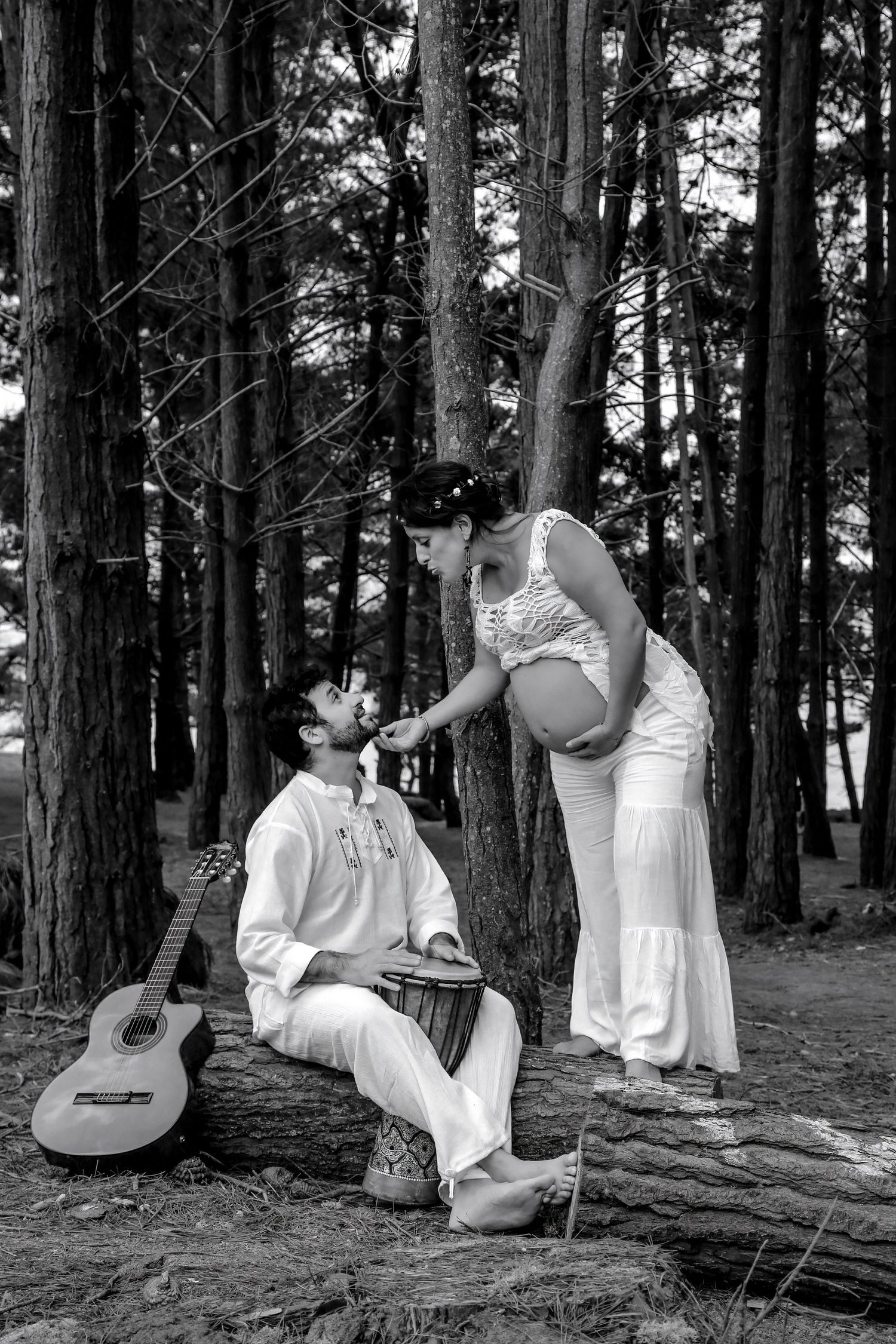 Black and white photo of a couple in white attire in a forest, man sitting with guitar and bongo drum.