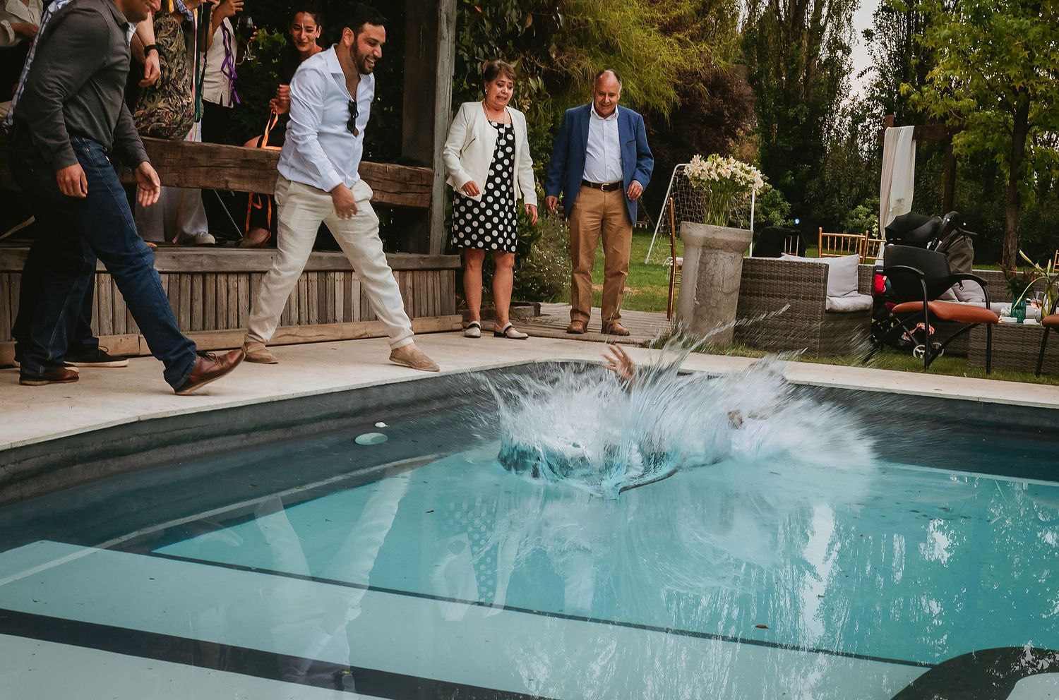Person jumping into outdoor pool at party, splashing water as guests in formal attire watch poolside.