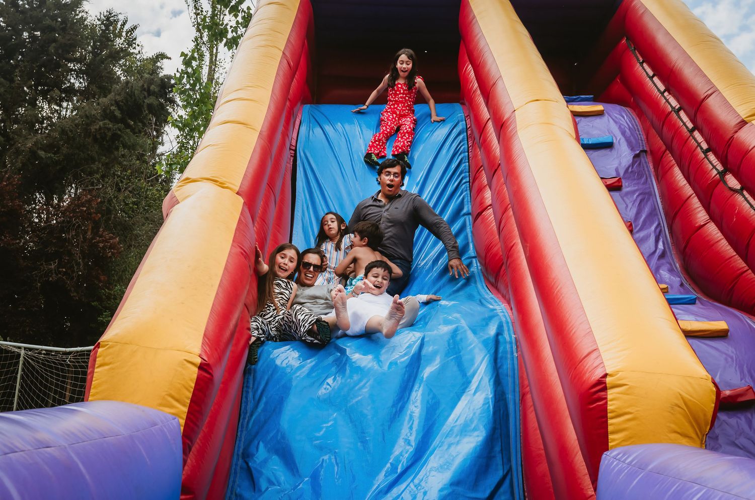 Children laughing and sliding down a colorful inflatable bounce house slide at an outdoor party.