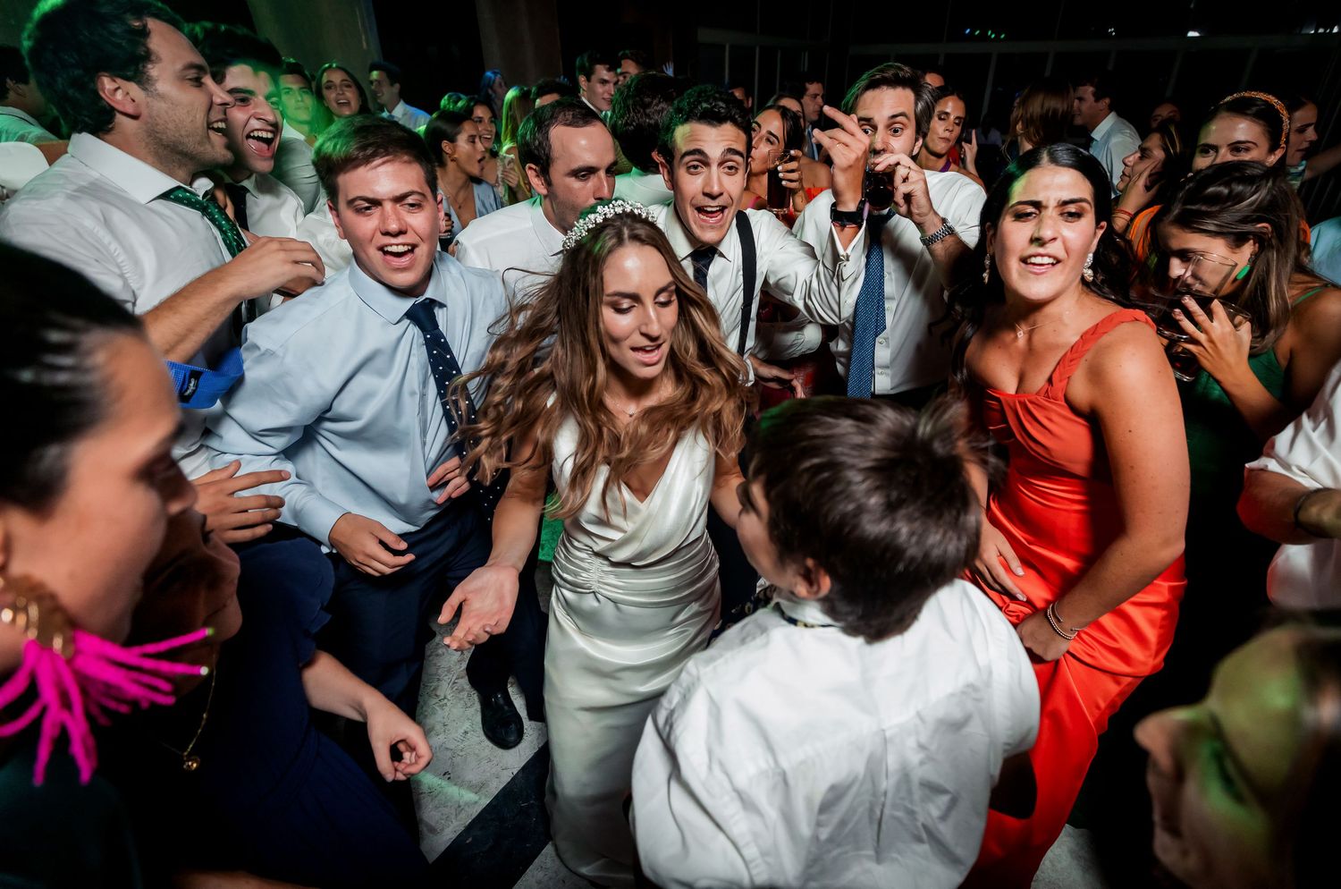 Smiling bride in white gown dancing joyfully surrounded by excited guests at energetic wedding reception.