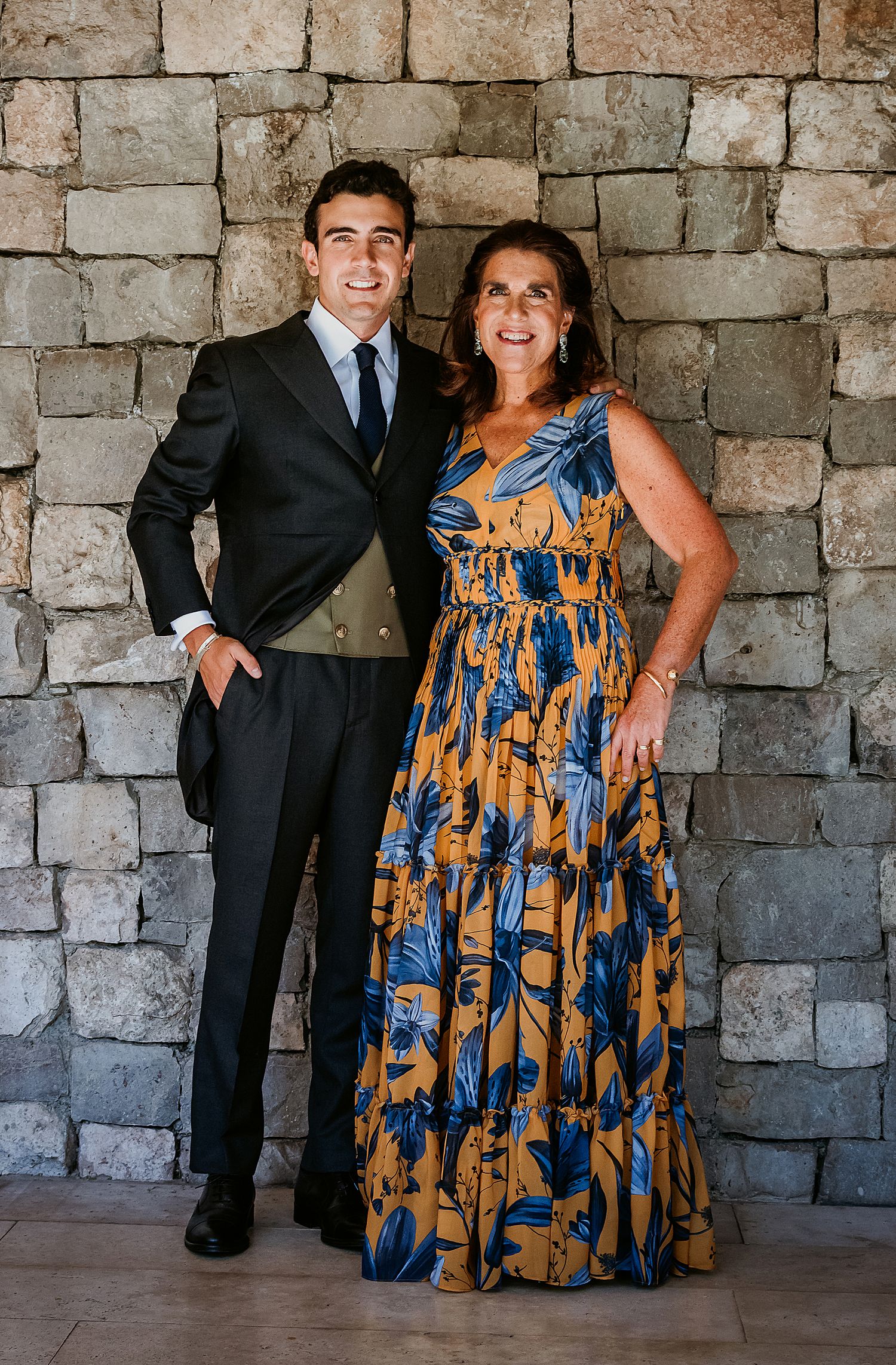 Smiling groom in black suit posing with woman in floral maxi dress against stone wall.