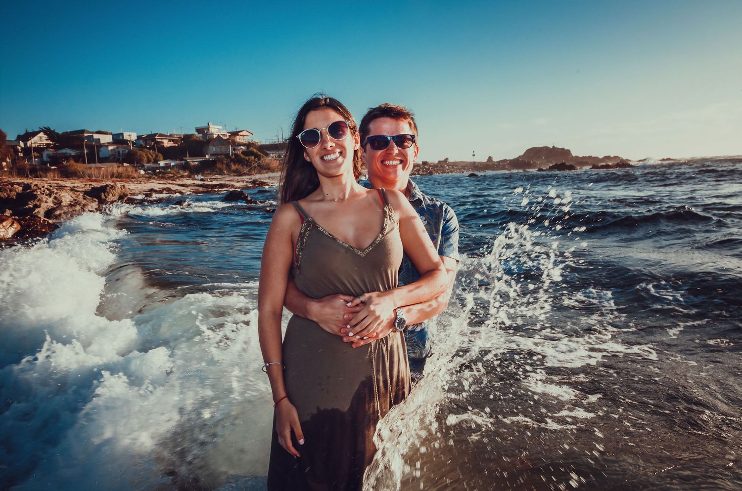 Happy couple smiling as waves crash behind them on a sunny beach shoreline.