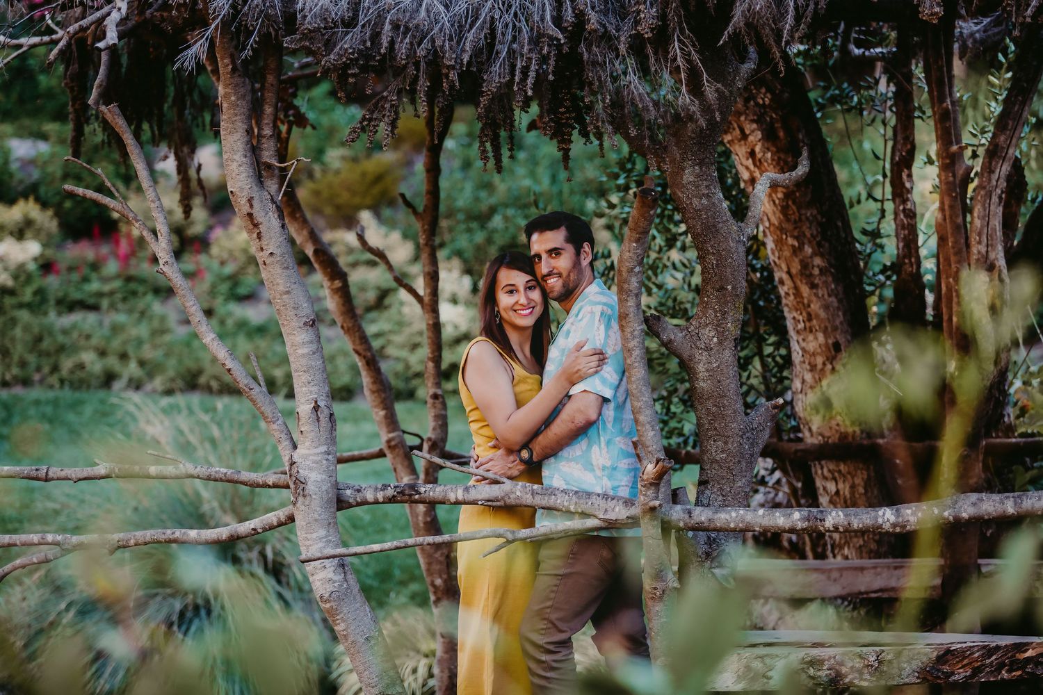 Couple cuddling on a rustic wooden bridge framed by trees and lush garden foliage.