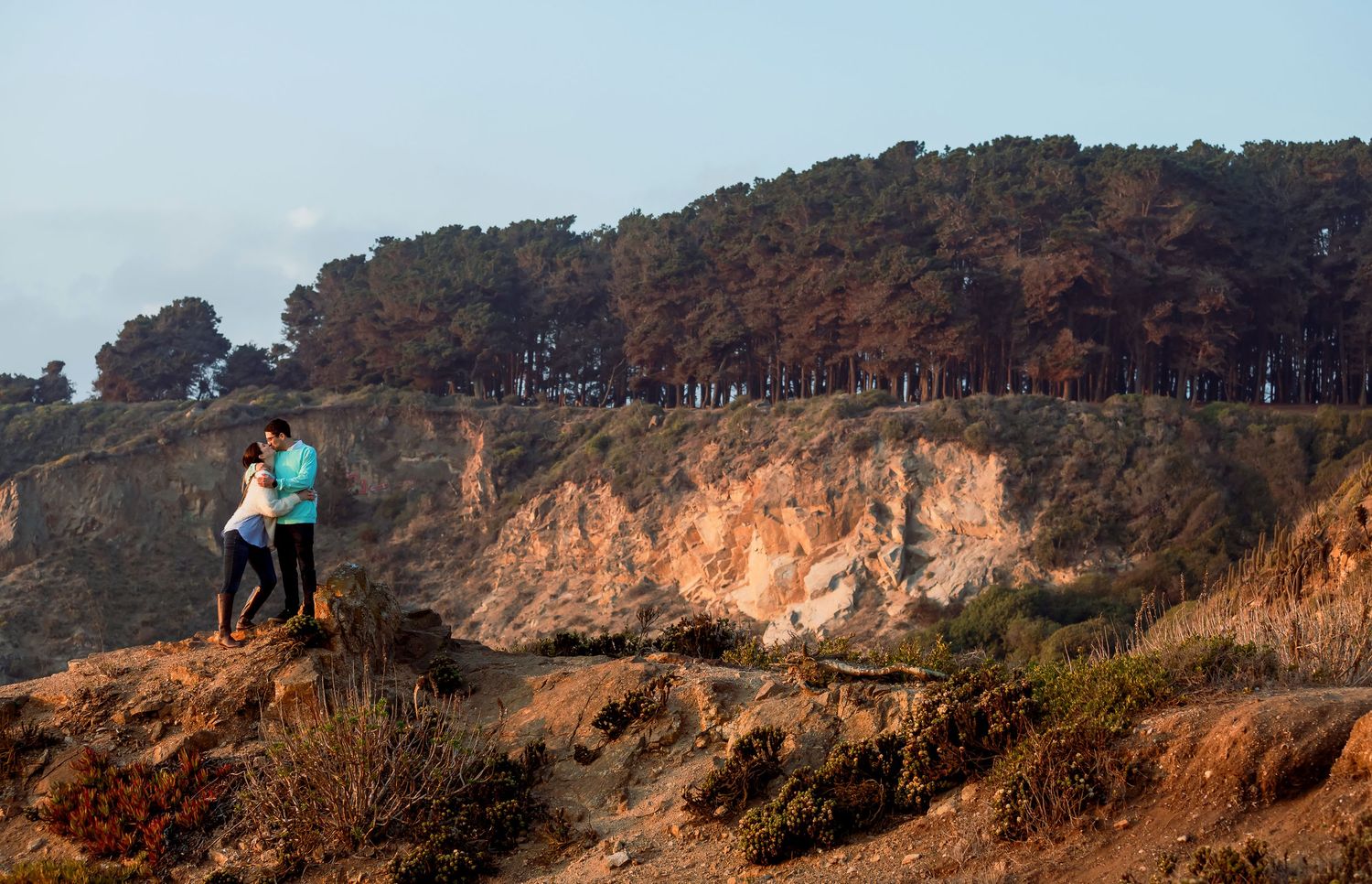 Couple standing together on dramatic coastal cliffs overlooking the ocean with pine trees in background.