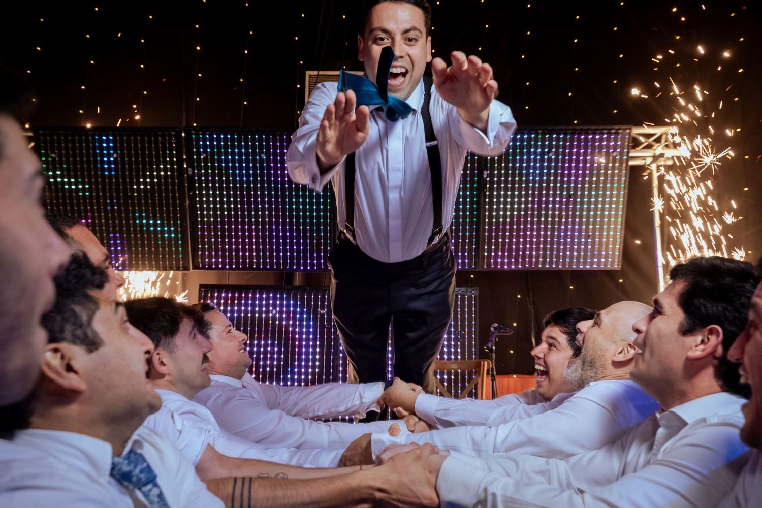 Groom crowd surfs over laughing groomsmen amid dazzling sparklers at a vibrant wedding party.