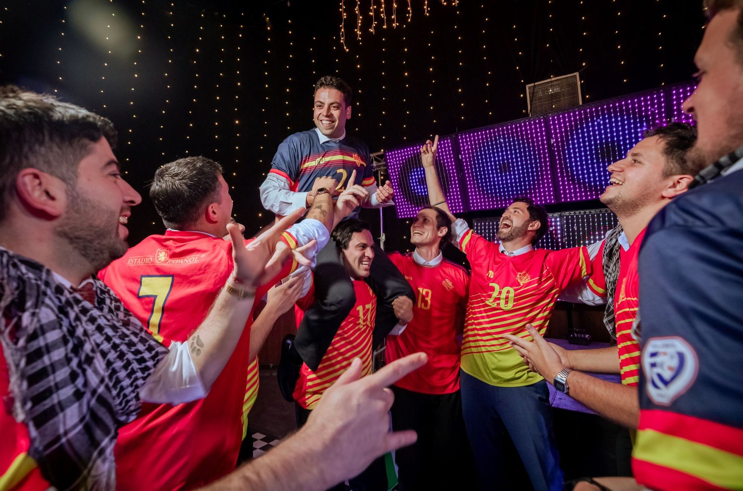 Groom lifted by cheering friends in soccer jerseys during a spirited wedding reception celebration.