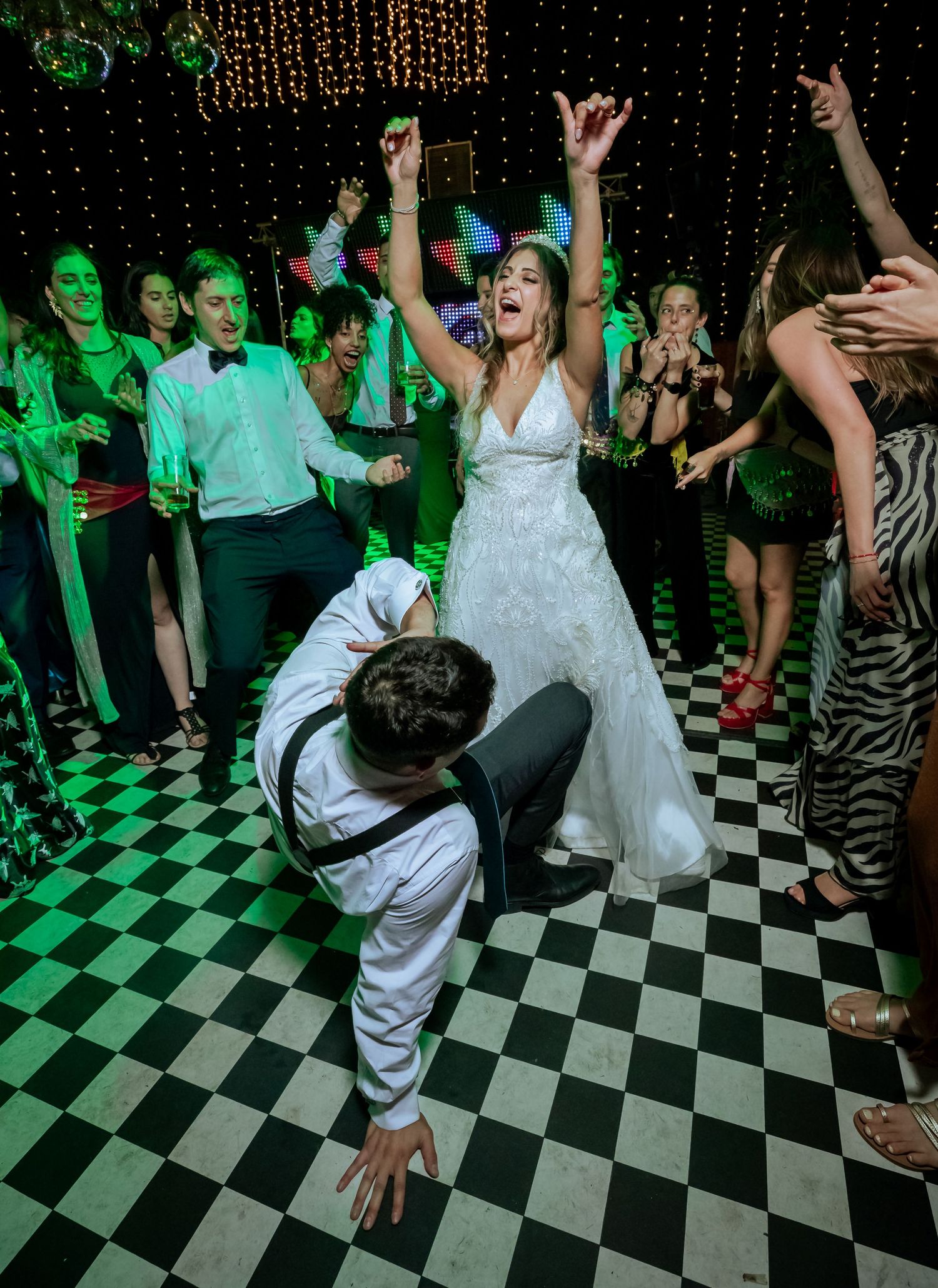 Bride raises her arms in celebration while groom bows on a black and white checkered wedding dance floor.