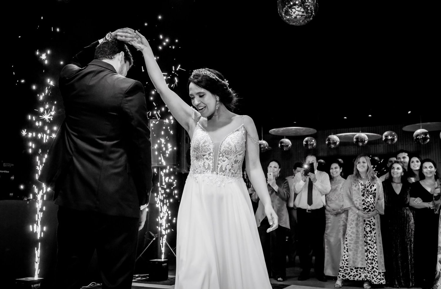 Groom spinning bride in white gown on dance floor surrounded by guests and sparkling fireworks.