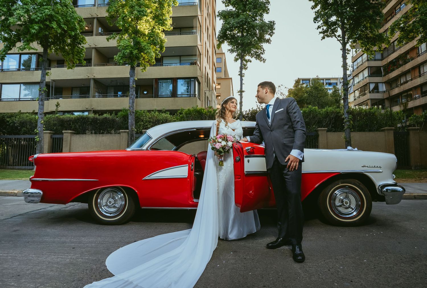 Wedding couple standing by a stunning red and white vintage classic car on a tree-lined urban street.