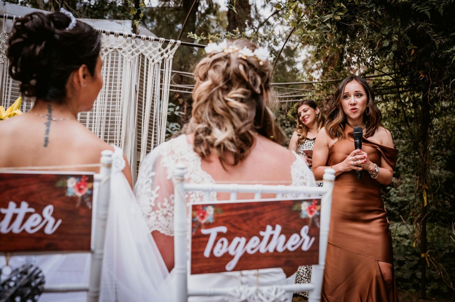 Officiant speaks at outdoor wedding ceremony as two brides sit in decorated Together chairs under macrame arch.