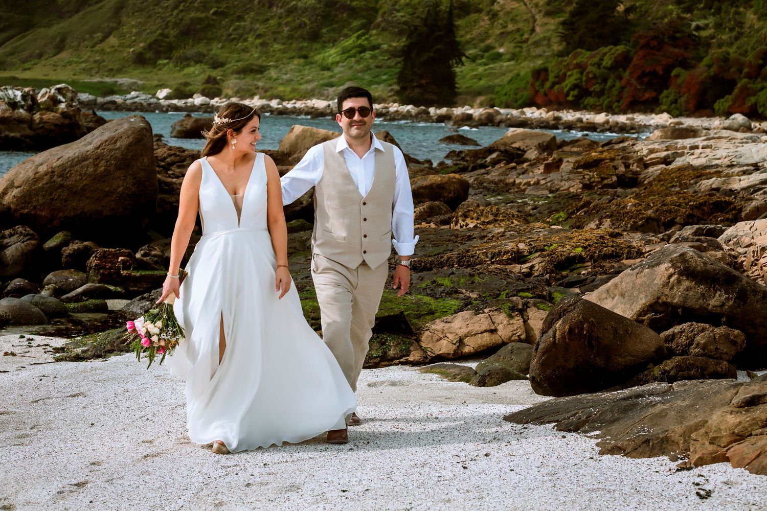 Newlyweds walking hand in hand along rocky shoreline, bride in flowing white gown, groom in tan suit.