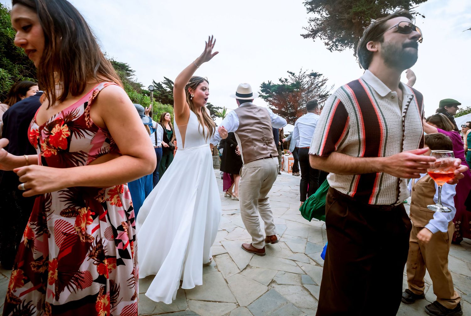 Bride in white gown dancing with wedding guests on a stone patio at outdoor reception.