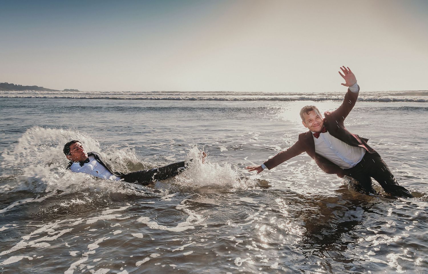 Two grooms in suits play in ocean waves, one falling back as the other reaches out on the beach.
