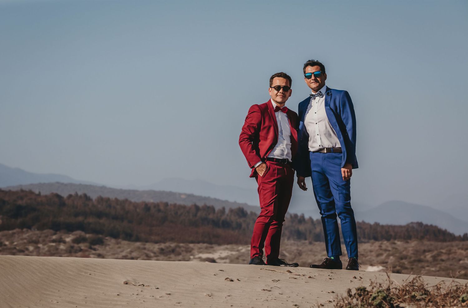Two grooms in red and blue suits standing together on sandy desert landscape under clear sky.