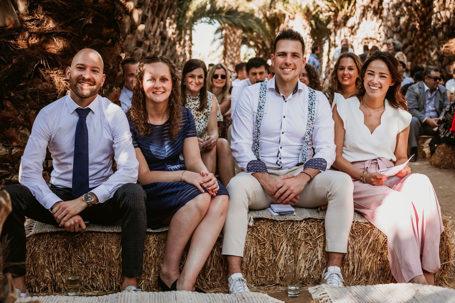 Four smiling wedding guests seated on hay bales at outdoor rustic palm tree wedding ceremony.