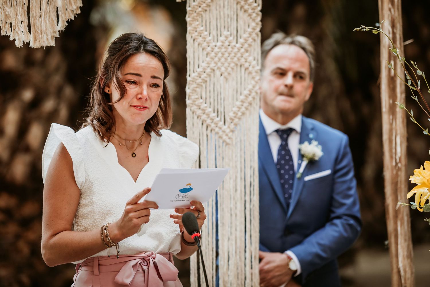 Emotional woman in white dress reads speech at outdoor wedding ceremony with macrame backdrop.