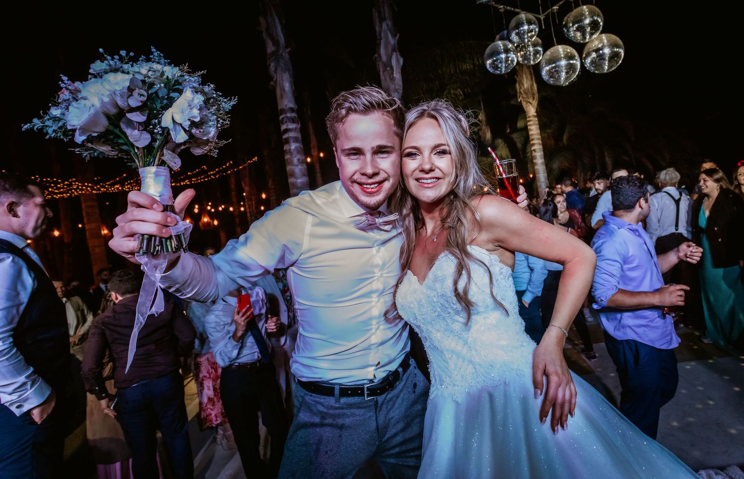 Happy couple poses with bouquet at outdoor wedding reception, smiling under string lights at night.