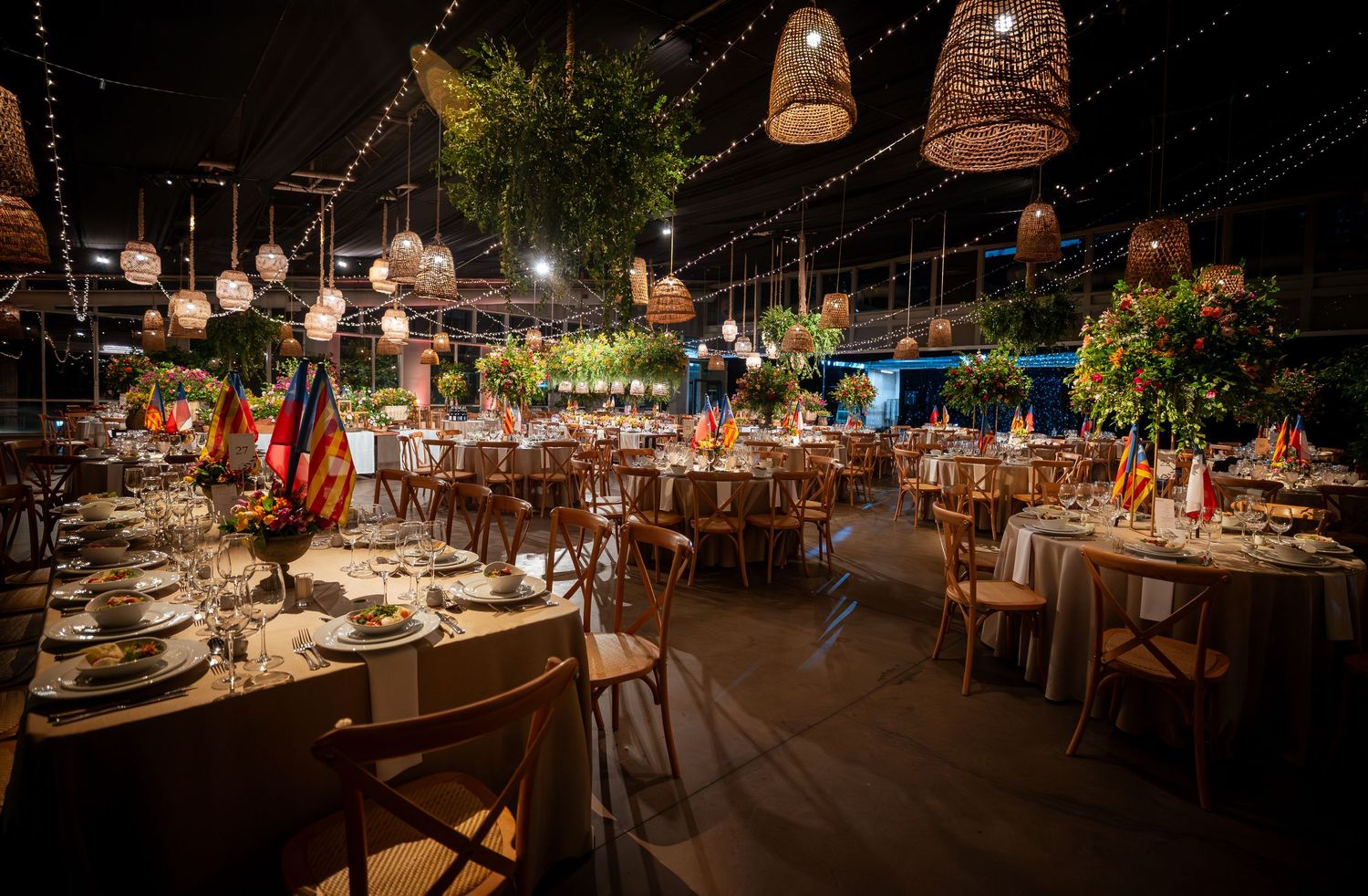Romantic wedding venue with wicker pendant lights, round tables, string lights, and tropical plants in evening setting.